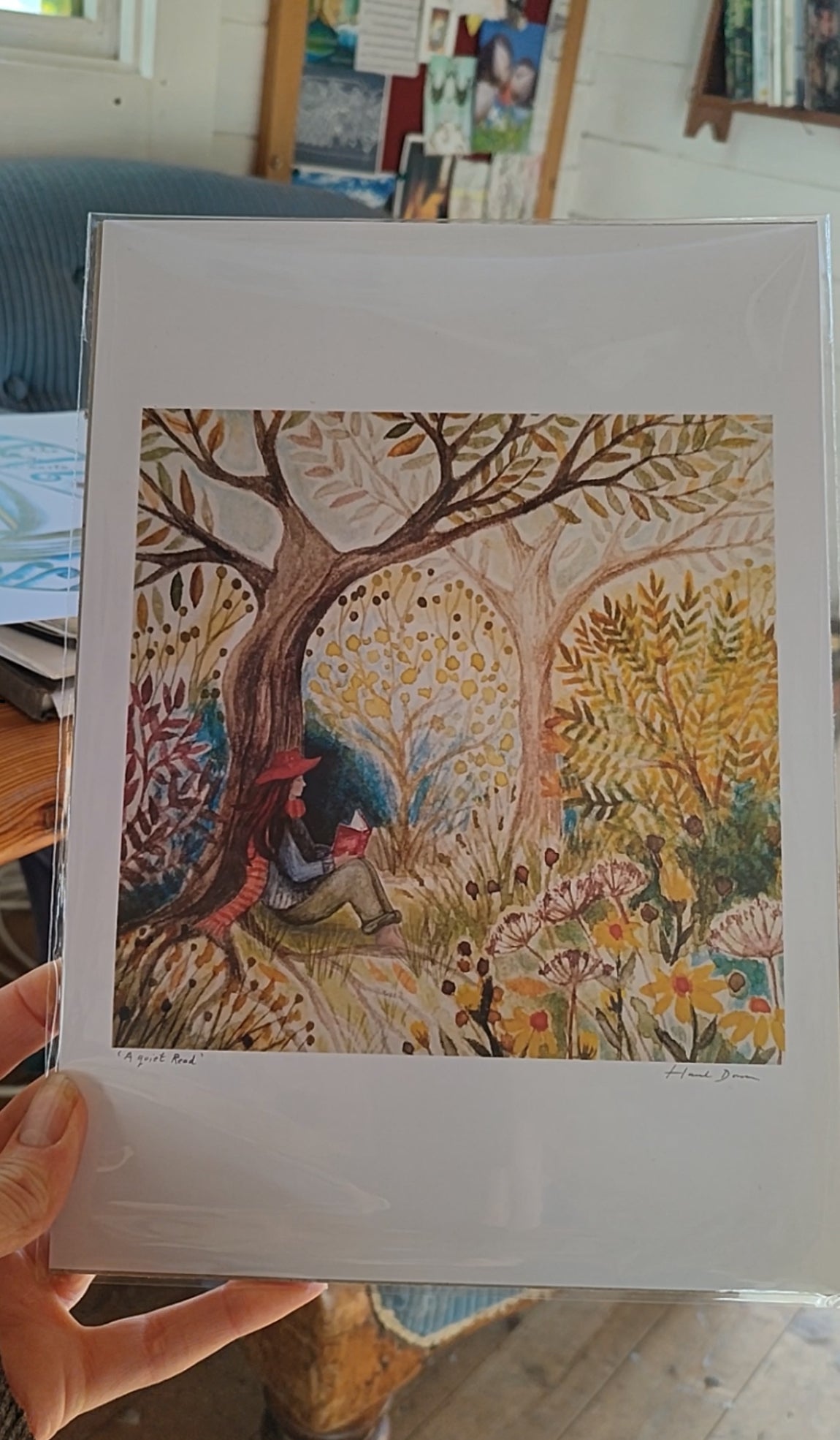 Artistic depiction of a person under a tree with colourful foliage, held in front of a blurred indoor background. Quiet reading spot in the woods, watercolour print, framed, slow-living art, mindful art, gifts for readers