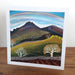 A square greeting card featuring a mountain landscape with trees and birds, printed on recycled paper with a recycled white paper envelope, placed on a wooden surface. Painting of a landscape with mountains, trees, and a blue sky. Mourne mountains art, hiker gift, Northern Ireland Artist Hannah Dorman, Autumn art 