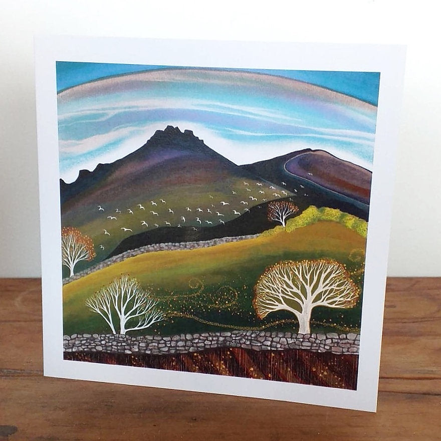 A square greeting card featuring a mountain landscape with trees and birds, printed on recycled paper with a recycled white paper envelope, placed on a wooden surface. Painting of a landscape with mountains, trees, and a blue sky. Mourne mountains art, hiker gift, Northern Ireland Artist Hannah Dorman, Autumn art 