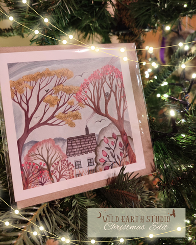 Card with a winter scene of trees and a house, placed on a Christmas tree with lights. Greeting card of a house surrounded by trees with autumn foliage. Cottage core autumn card with cosy cottage, owl in tree rosehips and birds in the sky, chimney smoke, slow living inspiration 