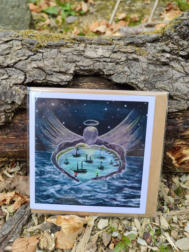 A square greeting card with a recycled paper envelope, featuring an artistic depiction of boats on water with a figure holding a halo above it, presented on a wooden surface with a compostable bag.