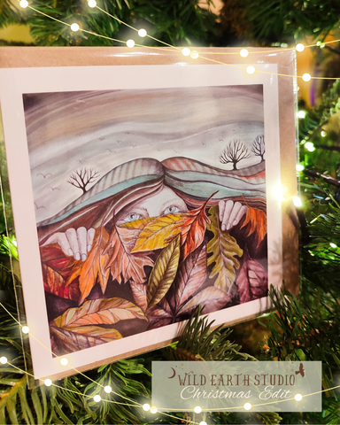 Illustration of a person with autumn leaves in front of a snowy landscape, framed and displayed on a tree with lights. Artistic depiction of a woman resting under ablanket of autumn leaves in front of a landscape, hair flowing into ploughed fields and trees, wintering card