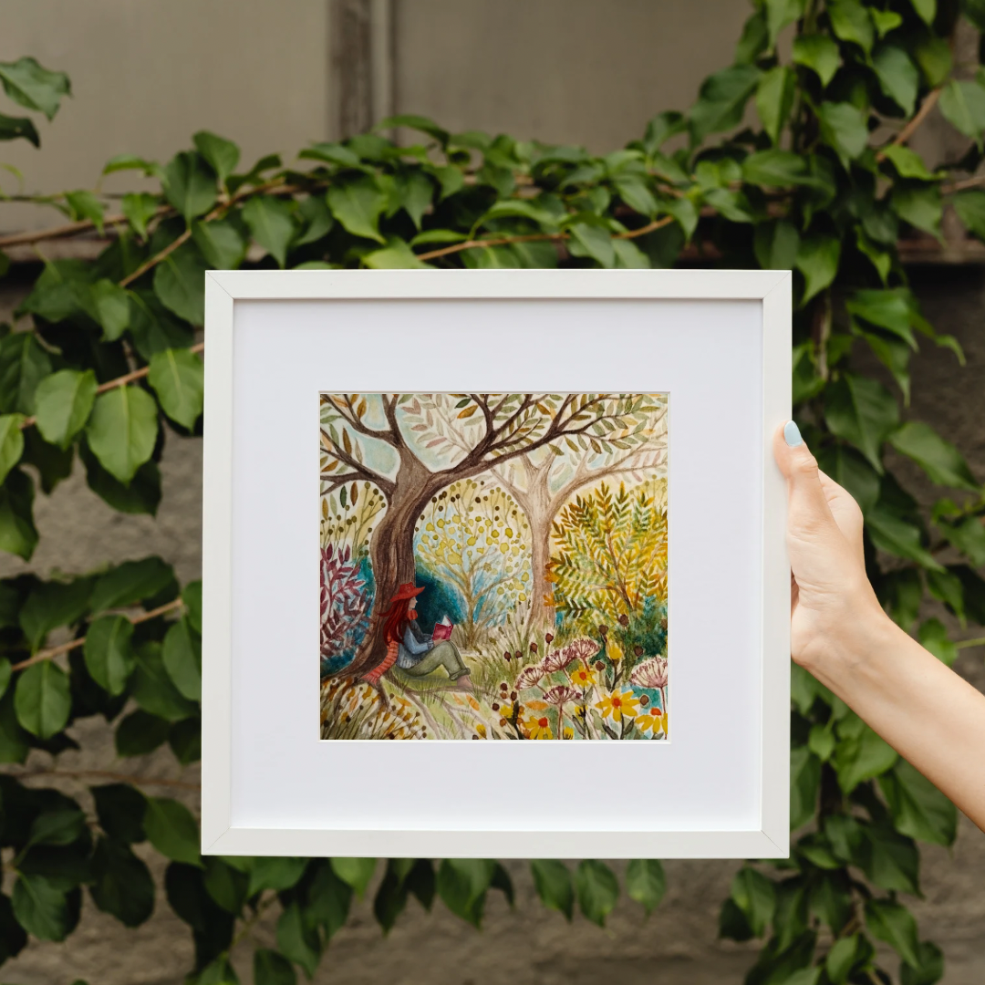 Framed artwork of a colourful forest scene held by a hand against a leafy background, Quiet reading spot in the woods, watercolour print, framed, slow-living art, mindful art, gifts for readers